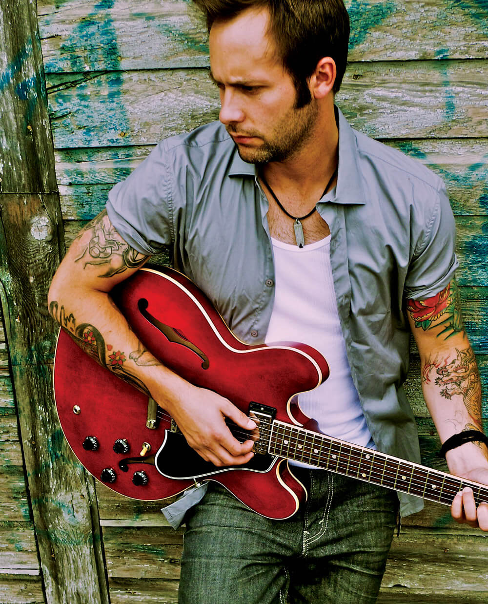 Dallas Smith wearing blue jeans, white t-shirt and a blue long-sleeved shirt over, leaning on a weathered wood wall playing a red electric acoustic guitar